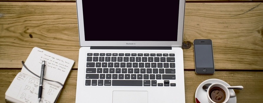 Otwarty laptop na drewnianym biurku, po jego lewej stronie leży długopis i zapisany notes, po prawej