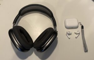 Na białym blacie dwie pary słuchawek. Pierwsze od lewej to duże nauszne słuchawki Apple Airpods Max w kolorze ciemno szarym. Charakterystyczne aluminiowe muszle i materiałowe poduszki nauszników oraz pałąk wypełniony drobną siateczką. Po prawej dwie pojedyncze słuchawki Airpods Pro w kolorze białym, a nad nimi białe błyszczące etui ładujące z zamontowanym paskiem na rękę.