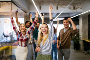 zdjęcie przedstawia grupę młodych osób, pozujących wspólne do zdjęcia w przestrzeni biurowej. Są uśmiechnięci i podnoszą ręce w geście zwycięstwa.