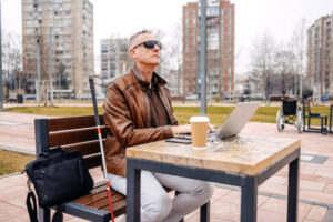 mężczyzna z niepełnosprawnością wzroku, siedzący na ławce w parku i pracujący na laptopie