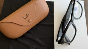 Jasnobrązowe etui na okulary z logo Ray-Ban. Obok etui leżą czarne okulary