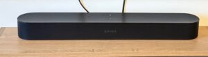 Czarny soundbar marki Sonos, ustawiony na drewnianej szafce pod białą ścianą. Z tyłu urządzenia widać wychodzące przewody. Soundbar ma zaokrąglone rogi i minimalistyczny design, a na jego froncie znajduje się logo "SONOS". Na górze obudowy widoczne są delikatnie podświetlone wskaźniki i przyciski dotykowe.