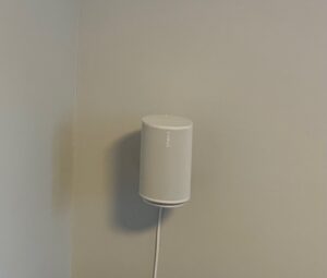 Biały głośnik Sonos Era 100 zamontowany pionowo na ścianie, z białym przewodem zwisającym w dół. Ściana jest gładka i pomalowana na jasnoszary kolor. Głośnik znajduje się w rogu pomieszczenia.