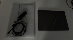 Na białym blacie ułożone obok siebie jasno-szare etui oraz niemal czarny zamknięty laptop Asus Zenbook S14. Na Etui leży niewielki czarny zasilacz ze zwiniętym, spiętym na rzep kablem do ładowania. Na pokrywie laptopa widoczny charakterystyczny wzór symbolizujący serię Zenbook.