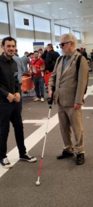 niewidomy mężczyzna, ubrany w brązowy garnitur, poruszający się z białą laską i detektorem. Stoi na korytarzu w przerwie konferencji.