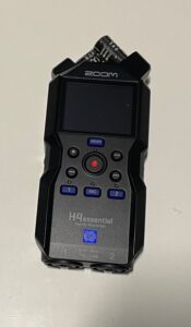 Rejestrator Zoom H4 Essential w kolorze czarnym o kanciastych kształtach, leżący na białym blacie. Ukazany frontem, w jego górnej części ukierunkowane pod lekkim kątem na prawo i lewo mikrofony. Pod nimi na froncie napis ZOOM i wyświetlacz. Na środku urządzenia oznaczony na czerwono przycisk nagrywania otoczony klawiszami nawigacyjnymi takimi jak stop, play/pauza, do przodu oraz do tyłu. Nad tą sekcją granatowy klawisz ustawiania czułości nagrywania, a pod nią trzy granatowe klawisze wyboru poszczególnych wejść oznaczone jako 1, 2 i Mic.